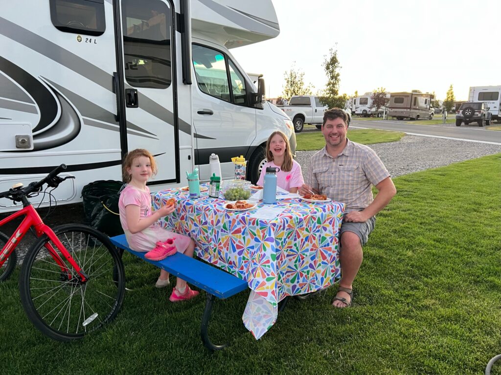 Family enjoying dinner outdoors next to an RV at a campground, highlighting RV rental lifestyle