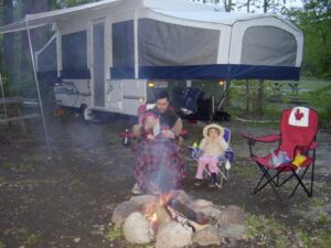 Family sitting by a campfire outside their pop-up camper at a campground.